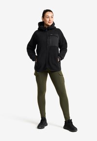 Svart fleecejacka med en dragkedjeförsedd bröstficka, tillsammans med olivgröna leggings med flera cargo-fickor och svarta sneakers.