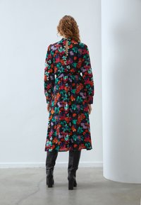 Mønstret langermet kjole i svart med livlige røde, grønne og lilla blomster; har en innsnørt midje og en nøkkelhullsdetalj bak.