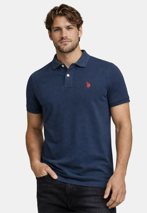 Mann mit kurzen lockigen Haaren, der ein marineblaues Poloshirt mit einem roten gestickten Logo trägt und mit einer Hand in der Tasche dasteht.