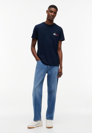 Mann iført en marineblå Tommy Jeans-T-skjorte, lyseblå jeans med rette ben og hvite joggesko, stående med en hånd i lommen.