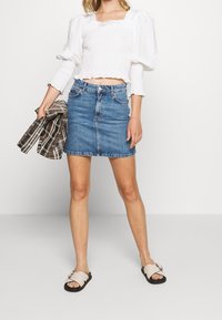 Valkoinen smokkaukseen tehty puffihihainen paita, farkkuinen mini-hame ja vaaleat slaidi-sandaalit. Malli pitää kädessään ruudullista takkia. Siisti, rento tyyli.