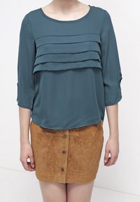 Blouse teal avec des volants superposés à l'avant et des manches trois-quarts, associée à une jupe mini en daim beige avec une fermeture à boutons à l'avant.