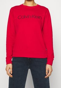 Czerwona bluza z prążkowanym okrągłym dekoltem, długimi rękawami i wyszywanym czarnym logo "Calvin Klein" na przodzie. Materiał z mieszanki bawełny.