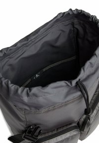 Zaino in tessuto grigio con apertura a coulisse, dotato di un interno liscio, tasca con zip e hardware per cinturini regolabili sui lati.