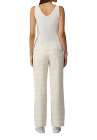 Vrouw in een mouwloze witte top met een V-rug, textuur beige wijde pijpenbroek en witte sneakers, staand met haar rug naar de camera tegen een witte achtergrond.