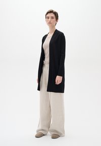 Zwart lange cardigan van zachte stof, gecombineerd met een beige top en lichtbeige wijde broek. Eenvoudig ontwerp zonder zichtbare patronen.