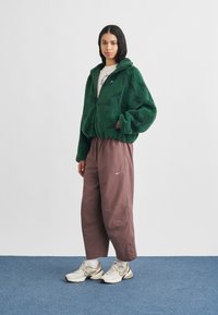 Πράσινο φλις μπουφάν με φερμουάρ και κουκούλα, σε συνδυασμό με φαρδιά καφέ παντελόνια και μπεζ αθλητικά παπούτσια. Απλός σχεδιασμός και απαλή υφή.