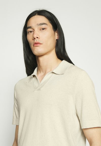 Jeune personne aux longs cheveux noirs portant une chemise beige à manches courtes avec col, regardant légèrement sur le côté, fond neutre.