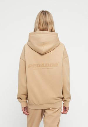 Beige Kapuzenpullover mit weicher Textur. Auf der Rückseite befindet sich ein Logo "PEGADOR" in einem dunkleren Farbton sowie gerippte Bündchen und Saum.