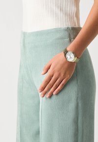 Pantalons en velours côtelé teal clair avec des côtes verticales, associés à une montre en ton doré avec un cadran blanc et des chiffres romains, portée à la taille.