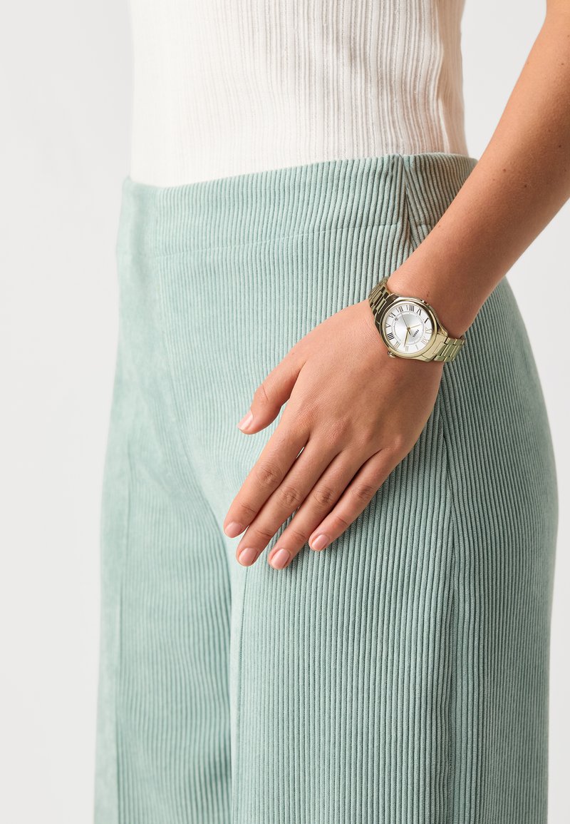 Pantalons en velours côtelé teal clair avec des côtes verticales, associés à une montre en ton doré avec un cadran blanc et des chiffres romains, portée à la taille.