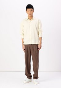 Light beige button-up shirt with long sleeves, paired with loose brown trousers and white sneakers, standing on a neutral background.