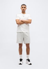 Νέος άνδρας με σταυρωμένα τα χέρια, φορώντας άσπρο t-shirt Puma, γκρι σορτς, άσπρες κάλτσες Puma και άσπρα αθλητικά παπούτσια, στέκεται μπροστά σε ένα απλό φόντο.