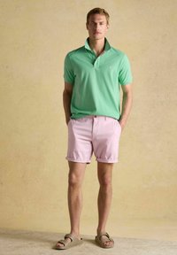 Zaļš polo krekls ar īsām piedurknēm, rozā šorti ar salocītām malām un brūnas sandales. Vienkāršs dizains un mīksta tekstūra.
