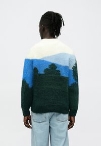 Mīksta adīta džempera ar ainavas dizainu zaļā, zilā un krēmkrāsā. Apaļa kakla līnija, garas piedurknes un brīvs piegriezums.