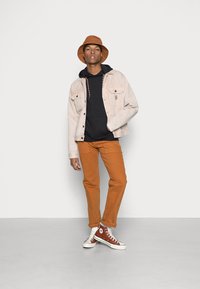 Lys beige denimjakke, sort sweatshirt, brændt orange bukser og brun bøttehat, parret med brune høje sneakers.