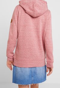 Person wearing a pink hooded sweatshirt and a blue denim skirt, shown from the back, standing against a plain light background.