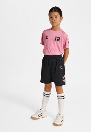 Jong meisje staat met handen achter de rug, draagt een roze sportshirt met nummer 12, zwarte korte broek, witte kniekousen en witte sneakers.