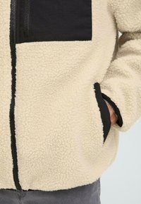 Cremefarbene Fleecejacke mit schwarzen Akzenten, ausgestattet mit einem Reißverschluss vorne, einer Brusttasche und Seitentaschen; strukturiertes, weiches Material.
