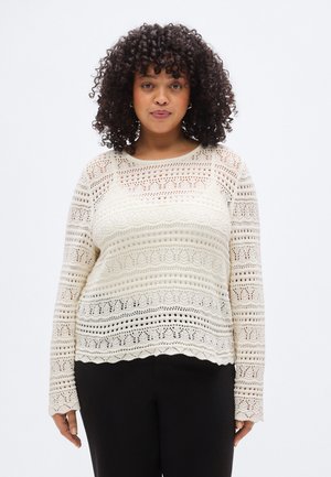 Cremefarvet blonderstrikket sweater med lange ærmer, der har et tekstureret mønster med hulstrik og bølget kant, kombineret med mørke bukser.