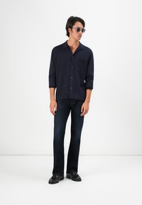 Marineblaues Hemd mit Knöpfen, einer Brusttasche und hochgerollten langen Ärmeln, kombiniert mit dunkelblauen geraden Jeans und schwarzen Slippern.