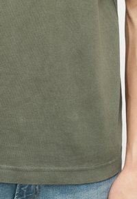 Κοντινό πλάνο στο τελείωμα ενός βαμβακερού T-shirt ελιάς, σε συνδυασμό με ανοιχτόχρωμο μπλε τζιν, με ορατό βραχίονα στη δεξιά πλευρά.