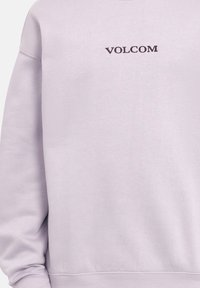 Sudadera de color púrpura claro con puños y dobladillo acanalados. Presenta un logo bordado en oscuro de "VOLCOM" en la parte delantera. Textura suave y lisa.