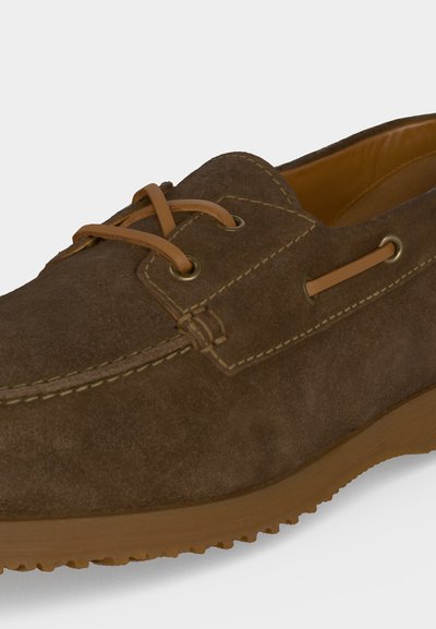 Mocassin en daim marron avec lacets et surpiqûres en cuir beige, doté d’une semelle en caoutchouc crantée pour une bonne adhérence.