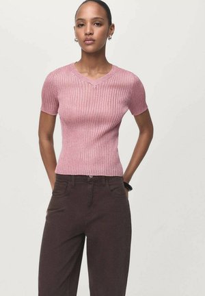 Ribbed short-sleeve top in light pink with a subtle V-neck. Fabric appears soft with a slight sheen. Paired with dark brown pants.