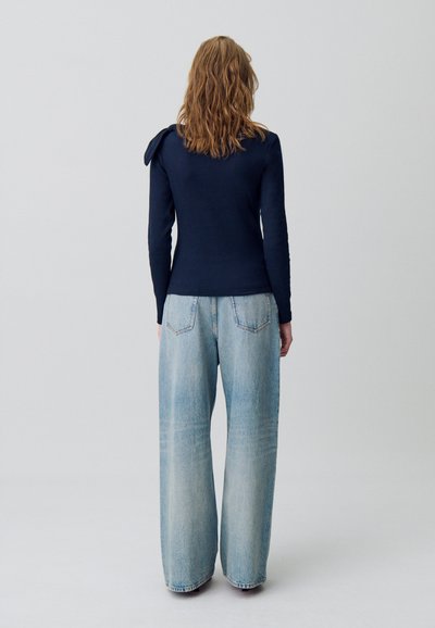 Maglietta a maniche lunghe blu navy con dettaglio sulle spalle abbinata a jeans in denim chiaro a gamba larga. Morbido al tatto e vestibilità rilassata. Sfondo semplice.