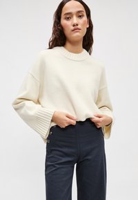 Πλεκτό πουλόβερ κρεμ χρώματος με ριμπ γιακά και φαρδιά μανίκια, συνδυασμένο με σκούρο παντελόνι ψηλόμεσο με διακοσμητικά κουμπιά.