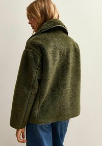 Blondyn w za dużej, oliwkowozielonej polarowej kurtce z wysokim kołnierzem, stojący na tle gładkiej, jasnej ściany.