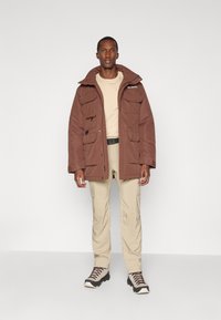 Veste isolée marron avec plusieurs poches, un col montant et une fermeture éclair. Associée à un pantalon cargo beige et des chaussures de randonnée.