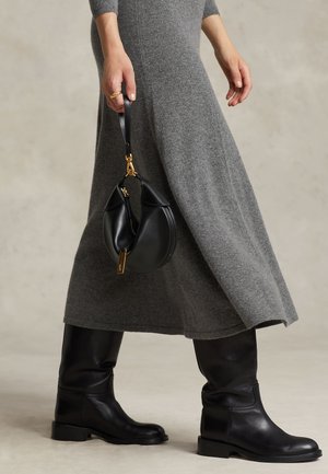 Isik kannab halli kudumist kleiti ja musti nahast põlviksaapaid, hoides käes väikest musta käekotti, millel on kuldsed detailid rihmal.