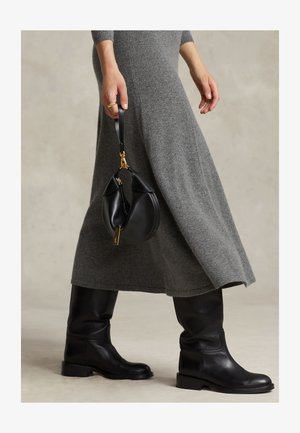 Isik kannab halli kudumist kleiti ja musti nahast põlviksaapaid, hoides käes väikest musta käekotti, millel on kuldsed detailid rihmal.