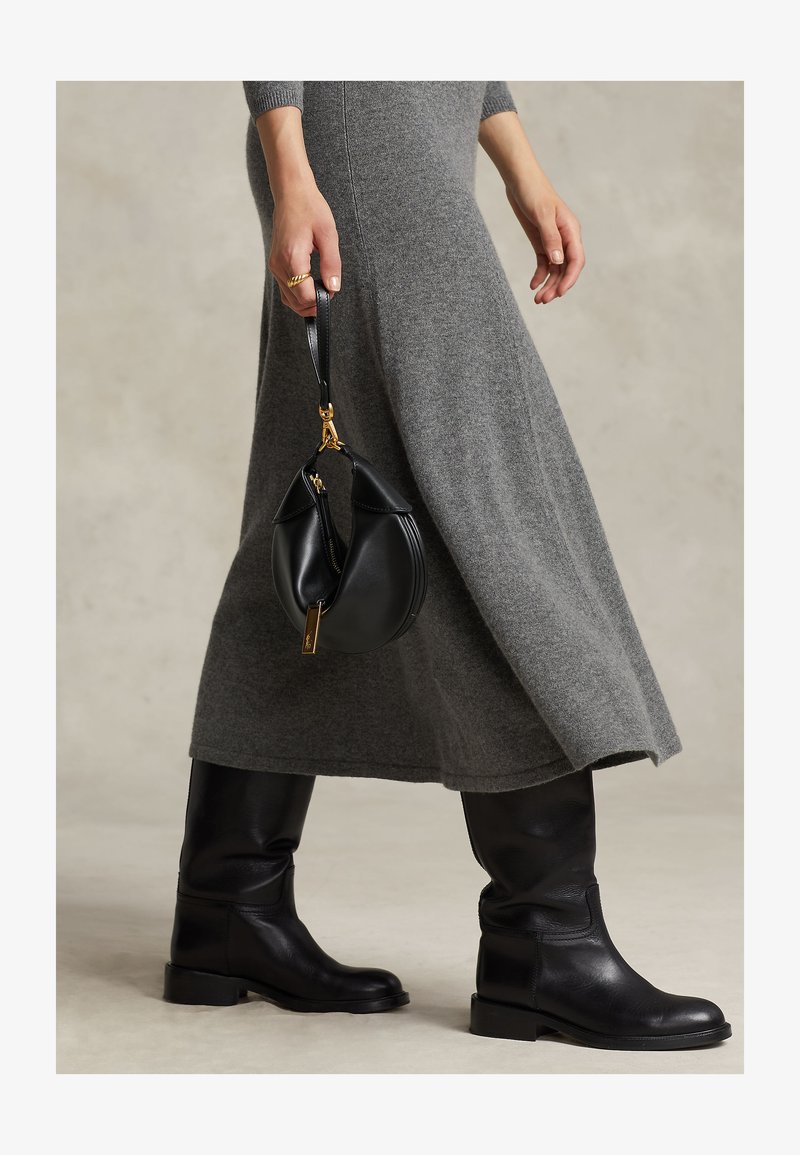 Osoba nosiaca sivé pletené šaty a čierne kožené čižmy po kolená, drží malú čiernu kabelku so zlatými kovaniami na popruhu.