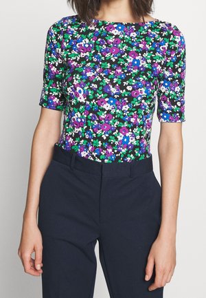 Femme portant un haut à manches courtes à motifs floraux bleu, violet, vert et blanc, associé à un pantalon noir taille haute, vu des épaules aux hanches.