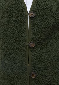 Veste en polaire texturée vert foncé avec trois grands boutons marron fermés sur le devant par-dessus une chemise blanche.