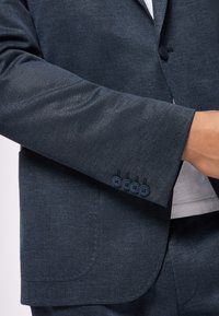 Dunkelgrauer Blazer mit feiner Textur, ausgestattet mit einem Ein-Knopf-Verschluss, drei Knopfapplikationen an den Ärmeln und einer strukturierten Passform.