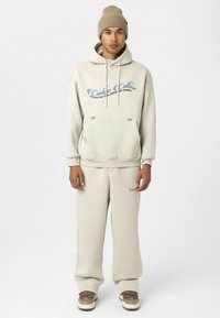 Crème-kleurige hoodie en sweatpants set met blauwe tekst en logo. Zachte stof, normale pasvorm en een capuchon met trekkoord. Draag het met beige sneakers.