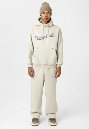 Crème-kleurige hoodie en sweatpants set met blauwe tekst en logo. Zachte stof, normale pasvorm en een capuchon met trekkoord. Draag het met beige sneakers.