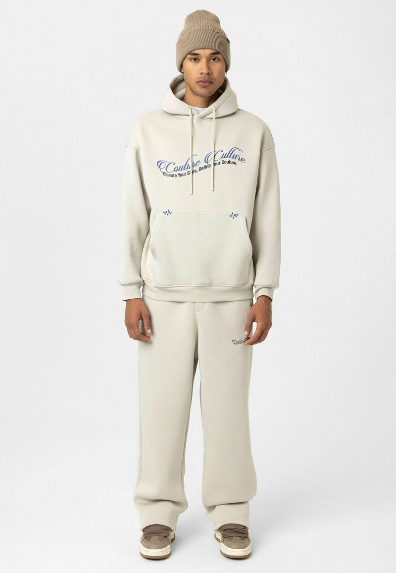 Crème-kleurige hoodie en sweatpants set met blauwe tekst en logo. Zachte stof, normale pasvorm en een capuchon met trekkoord. Draag het met beige sneakers.