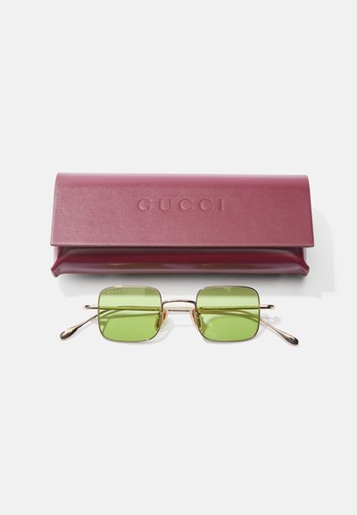 Gucci saules brilles ar kvadrātveida zaļiem lēcām, zelta metāla rāmjiem un melniem akcentiem. Komplektā iekļauta bordo ādas maciņš ar iegravētu "GUCCI."