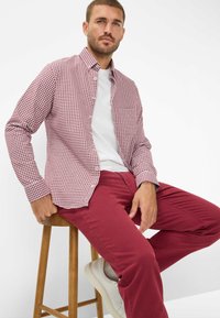 Camicia a maniche lunghe a quadri rossi e bianchi, indossata sopra una t-shirt bianca, abbinata a pantaloni rossi e sneakers bianche, seduto su uno sgabello di legno.
