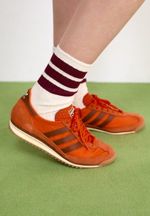 Des jambes portant des chaussettes blanches avec deux rayures bordeaux et des baskets Adidas orange sur un tapis vert.