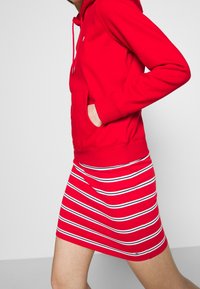 Roter Hoodie mit einer Fronttasche und Kordeln, kombiniert mit einem gestreiften roten Rock, der weiße und marineblaue Linien aufweist. Glatter Baumwollstoff.