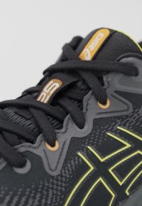 Gros plan sur une chaussure de course Asics noire avec des lacets noirs, des œillets orange et des accents jaunes du logo sur la tige en tissu tricoté.