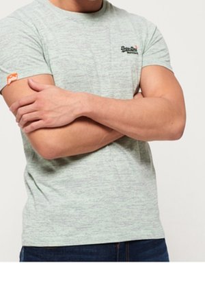 Mies, jolla on päällään vaaleanharmaa lyhythihainen t-paita, jossa vasemmassa rinnassa pieni logo, sekä siniset farkut, ristii kätensä.