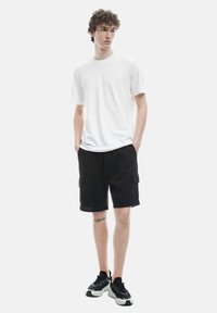 Camiseta blanca de cuello redondo, pantalones cortos cargo negros con bolsillos y zapatillas deportivas negras con suelas blancas. Atuendo casual en una figura de pie.