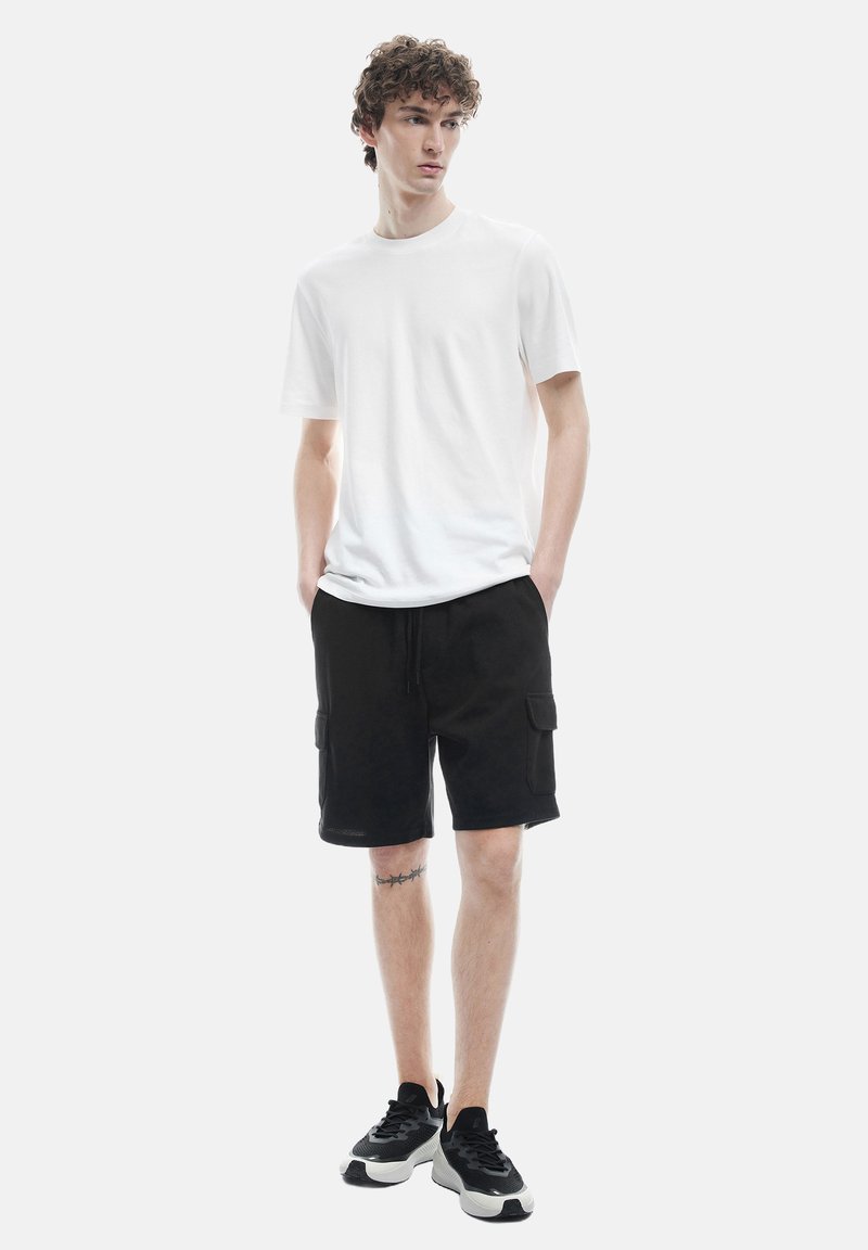 Camiseta blanca de cuello redondo, pantalones cortos cargo negros con bolsillos y zapatillas deportivas negras con suelas blancas. Atuendo casual en una figura de pie.